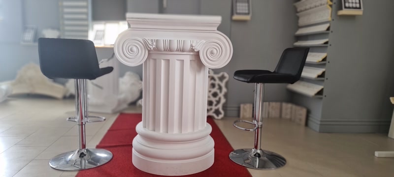 Kırmızı halı üzerinde sergilenen beyaz poliüretan iyonik sütun ve iki yanında siyah bar tabureleri.