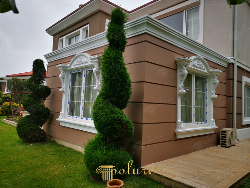 Kahverengi villa dış cephesinde beyaz poliüretan pencere sövesi, taç ve payanda uygulaması ile spiral bahçe ağaçları.